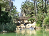 Через искусственный водопад перекинут деревянный мостик с аккуратными перилами. Мост соединяет два каменистых берега, наполненных яркой зеленью растений ...